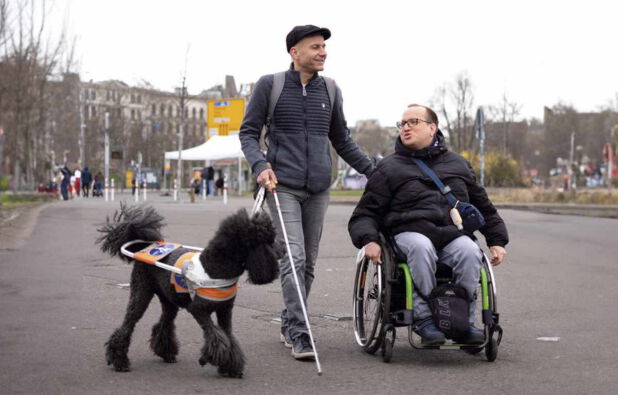 Das Foto zeigt zwei Männer, einer davon sitzt in einem Rollstuhl,  und einen Blindenführhund auf einem asphaltierten Weg.  Der Mann im Rollstuhl ist jüngeren Alters, hat dunkles, kurzes Haar und trägt eine Brille. Der andere befindet sich links daneben und hält sich am Griff des Rollstuhls fest. Dieser Mann trägt eine antrazitfarbene Jacke, eine Graue Jeanshose und eine Schiebermütze auf dem Kopf. Sein Gesicht ist dem anderen Mann zugewandt. In seiner rechten Hand hält er einen weißen Blindenlangstock und eine weiße Leine. An dieser befindet sich ein schwarzer Königspudel in seinem weißen Führgeschirr.