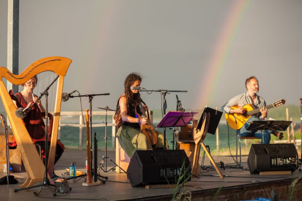Das Bild zeigt die drei Musiker sitzend unter freiem Himmel mit ihren Instrumenten auf einer Bühne. Sie halten eine Whistle, eine Fidel und eine Gitarre in den Händen. Auf der Bühne befinden sich außerdem eine Harfe, weitere Blasinstrumente sowie ein Harmonium. Im Hintergrund erstreckt sich ein stürmischer See. Ein doppelter Regenbogen erstrahlt über der stimmungsvollen Szenerie.