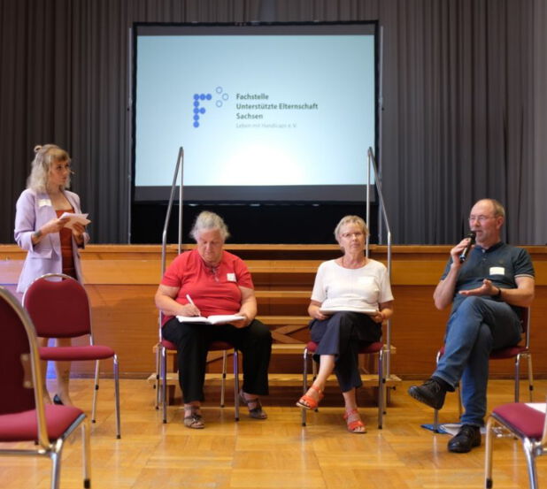 Die Fotografie zeigt 4 Personen. Links im Bild steht eine Junge Frau mit langen, blonden Haaren. Sie trägt ein fliederfarbenes Jackett über einem roten Oberteil. In beiden Händen hält sie in Brusthöhe Moderationskarten. Sie steht den drei anderen Personen zugewandt, welche nebeneinander auf Stühlen sitzen. Links sitzt Marion Michel. Sie trägt ein rotes T-Shirt und eine dunkle Hose. Sie blickt konzentriert. Rechts daneben sitzt eine Frau fortgeschrittenen Alters mit kurzen, blonden Haaren. Sie trägt ein weißes T-Shirt zu einer schwarzen Hose und eine Brille im Gesicht. Ganz rechts im Bild sitzt ein Mann mittleren Alters. Er trägt ein schwarzes T-Shirt zu einer blauen Jeans und hat eine Brille auf. Sein Mund ist leicht geöffnet. Er hält ein Mikrofon in der rechten Hand und gestikuliert mit der Linken. Im Hintergrund auf einer erhöhten Bühne befindet sich eine Projektionsfläche, auf dieser ist das Logo der Fachstelle Unterstützte Elternschaft abgebildet.