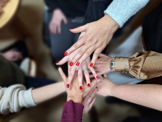 Im Zentrum des Fotos sind 7 übereinandergelegte Frauenhände mit unterschiedlichen Hauttönen abgebildet. Die Fingernägel der beiden oberen Hände tragen glänzend dunkelroten Nagellack.